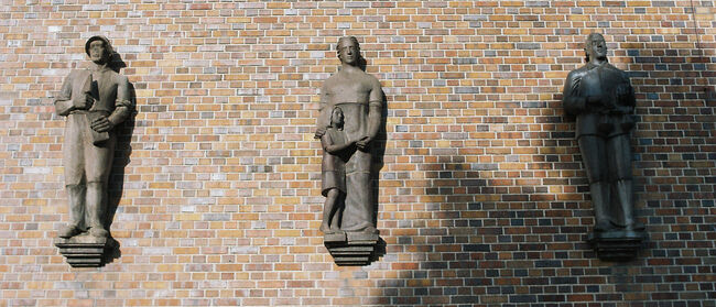Brick wall illuminated by the sun, on which three half-relief figures in the style of socialist realism are mounted on pedestals. The figures represent forms of work: on the left, a male bricklayer with a brick and trowel; in the center, a woman with a child; on the right, a lawyer with a weighing pan. The shadow of a tree falls across the lower right quarter of the picture and covers the figure on the right. 