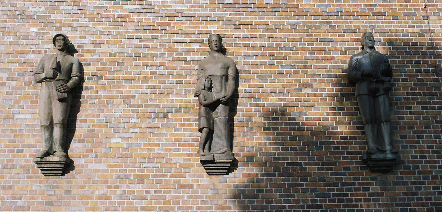 Brick wall illuminated by the sun, on which three half-relief figures in the style of socialist realism are mounted on pedestals. The figures represent forms of work: on the left, a male bricklayer with a brick and trowel; in the center, a woman with a child; on the right, a lawyer with a weighing pan. The shadow of a tree falls across the lower right quarter of the picture and covers the figure on the right.  (opens enlarged image)